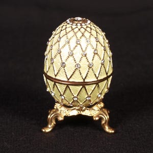 May include: A decorative egg-shaped trinket box with a pale yellow enamel finish and a diamond pattern. The egg is adorned with small, sparkling crystals and sits on a gold-colored, ornate stand.