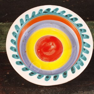 May include: A colorful ceramic plate with concentric circles in red, yellow, purple, and orange. The plate has a white rim with a teal leaf design. The plate is on a dark wooden surface.