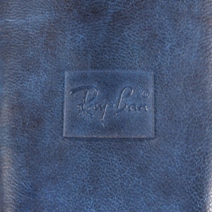 Puede incluir: Primer plano de un estuche de cuero azul con el logotipo de Ray-Ban en relieve. El estuche tiene un aspecto texturizado y un cierre de cremallera. Se ven detalles blancos y azules en los laterales.