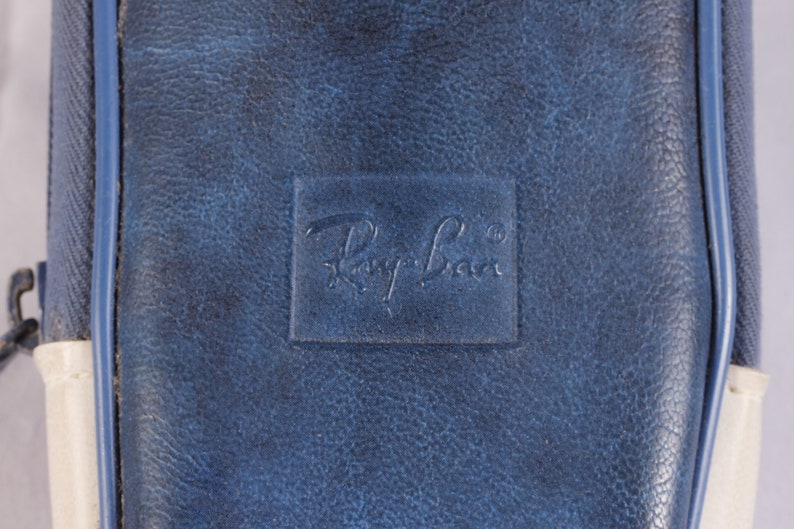 Puede incluir: Primer plano de un estuche de cuero azul con el logotipo de Ray-Ban en relieve. El estuche tiene un aspecto texturizado y un cierre de cremallera. Se ven detalles blancos y azules en los laterales.