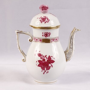 May include: A white porcelain coffee pot with a pink floral design and gold accents. The pot has a lid with a pink rose on top.