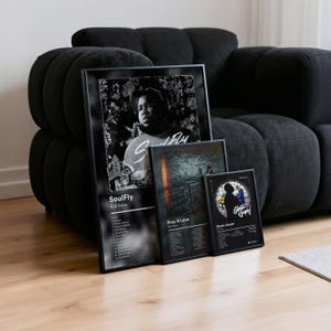 May include: Three framed music posters leaning against a black sofa. The largest poster features the text "SoulFly" and a portrait. The other two posters have album art and song lists. All posters are black and white.