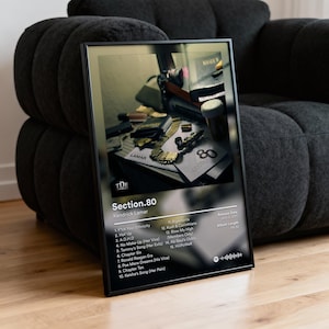 May include: Black framed poster featuring the album cover for Kendrick Lamar's "Section.80." The image within the frame shows a still life with gold jewelry and other objects. The album title and tracklist are visible on the poster.