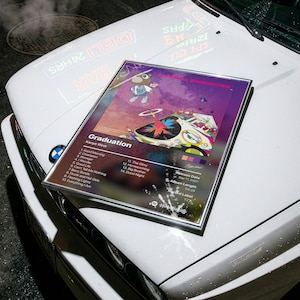 Peut inclure: Une affiche encadrée de l'album Graduation de Kanye West, présentant l'illustration colorée de Takashi Murakami, une liste de 14 titres, la date de sortie du 11 septembre 2007, la durée de 54:29, le label YZY et un code Spotify en bas.