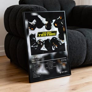 Puede incluir: Un póster enmarcado con la portada del álbum "Testing" de A$AP Rocky. El póster, con marco negro, muestra el título del álbum y la lista de canciones. La obra de arte presenta una imagen en escala de grises con el título en amarillo.