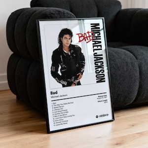 May include: Framed poster featuring Michael Jackson's album "Bad." The image shows Jackson in a black jacket. The poster includes the album title, tracklist, and release details. The word "Bad" is written in red. The poster is in a black frame.