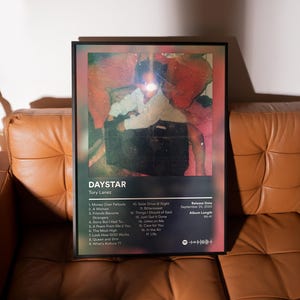 May include: A framed poster featuring the album "DAYSTAR" by Tory Lanez. The artwork includes a faded photograph of a person, with a tracklist and release details. The poster is displayed on a brown leather couch.