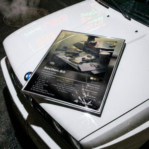 May include: A framed album cover for "Section.80" by Kendrick Lamar, resting on the hood of a white car. The album art features a grayscale image with text listing song titles and release details. The car's hood reflects neon signs.