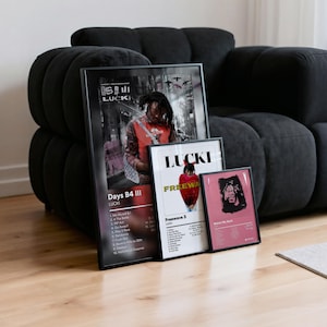 May include: Three framed art prints featuring the artist Lucki. The largest print displays the album cover for "Days B4 III," while the other two showcase "Freewave 3" and "Watch My Back." The prints are in black frames, leaning against a black sofa.