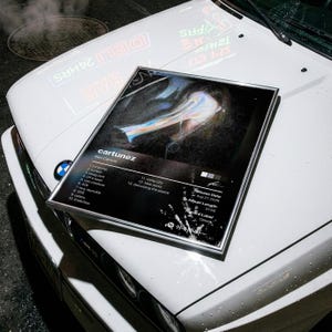 Peut inclure: Une affiche rectangulaire encadrée présentant la pochette de l'album Cartunez de Ken Carson. Le design comprend une image centrale sombre et abstraite, une liste de 13 titres, la date de sortie, la durée, le label et un code Spotify.