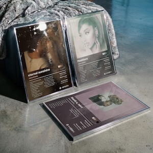 Könnte beinhalten: Drei gerahmte Albumkunstdrucke mit Ariana Grande. Die beiden oberen Rahmen zeigen die Alben "eternal sunshine" und "Positions", während der untere Rahmen "thank u, next" zeigt. Jeder Rahmen hat einen silbernen Rand.