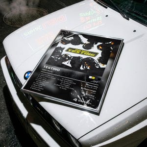 Puede incluir: Una portada de álbum enmarcada para el álbum "Testing" de ASAP Rocky, apoyada en el capó de un coche blanco. La portada presenta una imagen en blanco y negro con el título del álbum y la lista de canciones. El capó del coche está mojado.