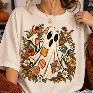 Puede incluir: Una camiseta de color crema con un gráfico de un fantasma blanco adornado con varios parches de tela coloridos. El fantasma está rodeado por una ilustración detallada de flores silvestres naranjas, amarillas y blancas, vegetación y trigo.