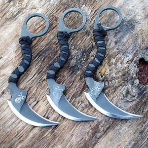 May include: Three gray karambit knives with curved blades and finger rings. The handles are wrapped in black cord. Each blade features a skull and crossbones design. The knives are displayed on a textured wooden surface.