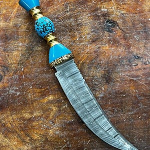 May include: A decorative knife with a patterned steel blade and a handle featuring turquoise-colored accents, gold-colored metal, and a textured blue bead. The knife is displayed on a wooden surface.