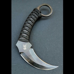 May include: A black karambit knife with a curved blade and a finger ring. The handle is wrapped in black cord. A skull and crossbones symbol is etched on the blade.
