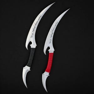 May include: Two curved silver blades with pointed tips and sharp edges. One handle is wrapped in black cord, and the other is wrapped in red cord. Each blade features an engraved inscription in a flowing script along the upper curved edge.