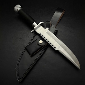 May include: A survival knife with a serrated blade and a black handle, resting in a black leather sheath. The knife has a silver blade and a silver metal cap on the handle. The sheath has a snap closure and a belt loop.