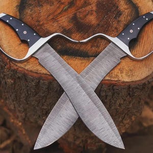 May include: Two crossed knives with curved handles and wide blades. The handles are black with white dots and a red accent. The blades have a textured pattern. The knives are displayed on a wooden surface.