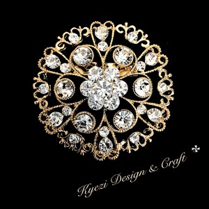 Peut inclure: Broche dorée ornée, avec un groupe central de cristaux clairs et étincelants, entouré de petits cristaux et de détails en filigrane. La broche a un motif floral et est posée sur un fond noir. Le texte "Kyezi Design & Craft" est visible.