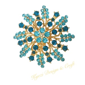 Op de afbeelding: Een gouden broche met een groot, ingewikkeld ontwerp met blauwe en turquoise strass steentjes. De broche heeft de vorm van een bloem met meerdere lagen bloemblaadjes.