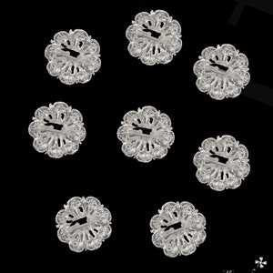 May include: Eight clear plastic flower-shaped buttons with a decorative design. The buttons have a hole in the centre and are arranged in a grid pattern on a black background.