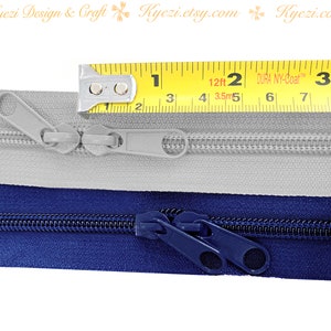 May include: Two zippers, one gray and one navy blue, are laid out side-by-side. A yellow measuring tape is placed above the zippers, showing a measurement of 2 inches.