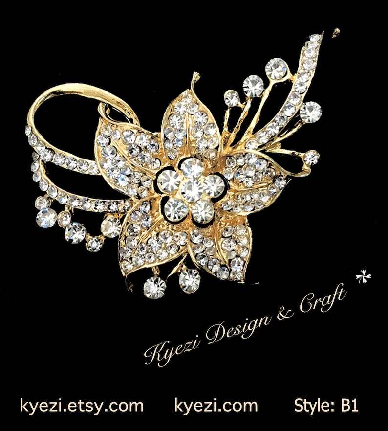 May include: A gold-coloured floral brooch adorned with clear crystal embellishments. The brooch showcases a flower design with multiple petals, surrounded by smaller crystals and a curved detail. The text "Kyezi Design & Craft" and website details are present.