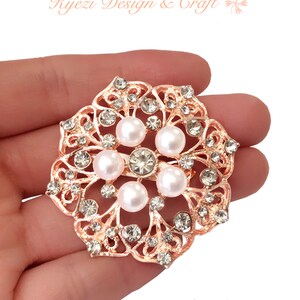 May include: Rose gold-toned brooch with a floral design, featuring faux pearls and clear crystals. The brooch has an openwork pattern and is round in shape. The text "Kyezi Design & Craft" and the website address are visible.