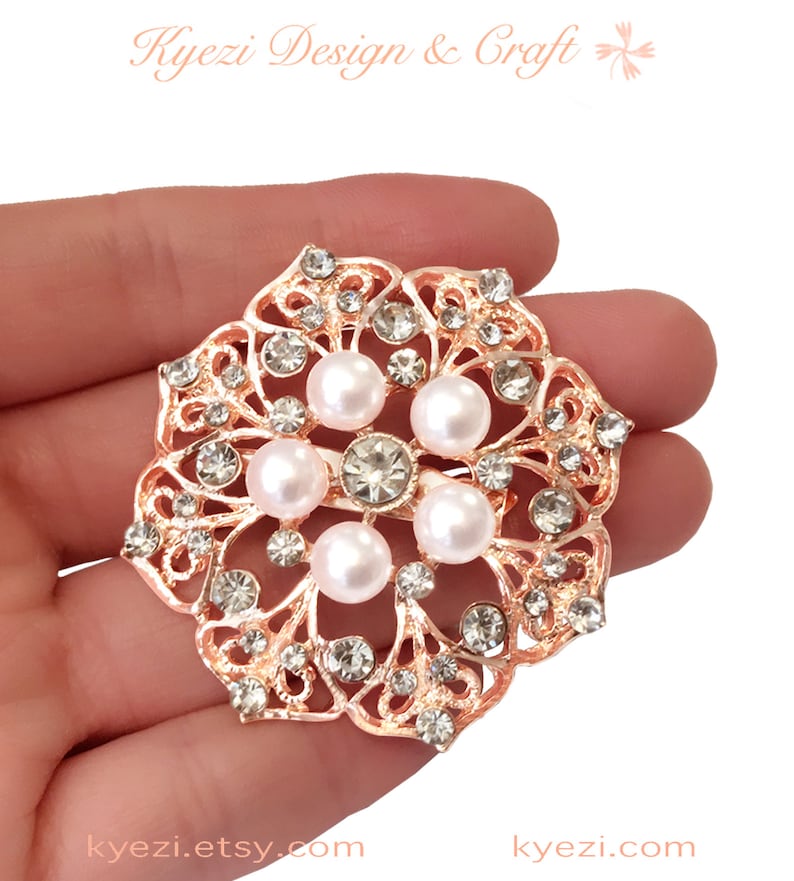 May include: Rose gold-toned brooch with a floral design, featuring faux pearls and clear crystals. The brooch has an openwork pattern and is round in shape. The text "Kyezi Design & Craft" and the website address are visible.