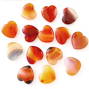 K&ouml;nnte beinhalten: Eine Sammlung herzf&ouml;rmiger Steine in Orange-, Rot- und Braunt&ouml;nen. Die polierten Steine weisen eine gl&auml;nzende Oberfl&auml;che und unterschiedliche Muster auf. Der Etsy-Shop-Name und die Website sind sichtbar.