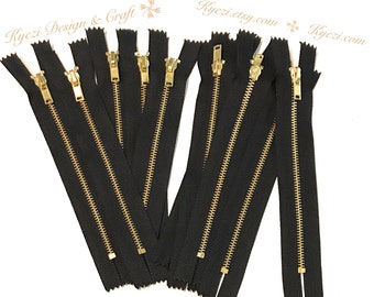 May include: Eight black zipper closures with gold metal sliders. The zippers are all the same length and are arranged in a row.