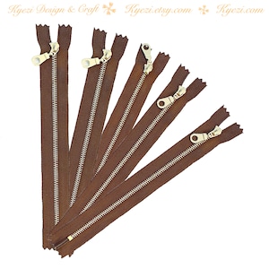 May include: A set of seven brown zippers with gold metal pulls. The zippers are arranged in a fan shape, with the longest zipper at the top and the shortest zipper at the bottom.