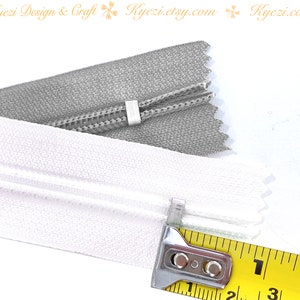 May include: Close-up of two zipper tapes, one white and one gray, measured with a yellow tape measure. The tape measure shows 1 inch.