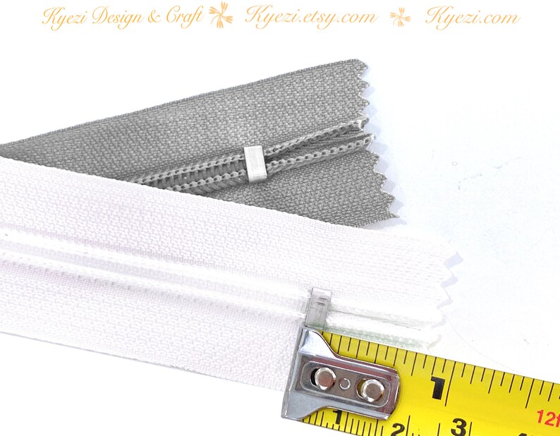 May include: Close-up of two zipper tapes, one white and one gray, measured with a yellow tape measure. The tape measure shows 1 inch.