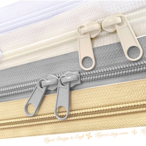 May include: Close-up of four different zipper tapes with metal sliders. The tapes are white, beige, gray, and tan. The sliders are silver and white.