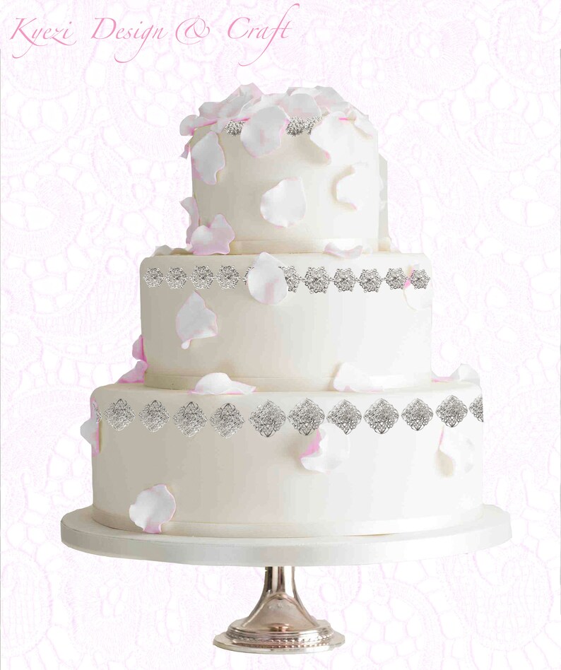 May include: A three-tiered white wedding cake, decorated with silver trim and scattered pink and white petals. The cake is on a silver stand. The text "Kyezi Design & Craft" is at the top.