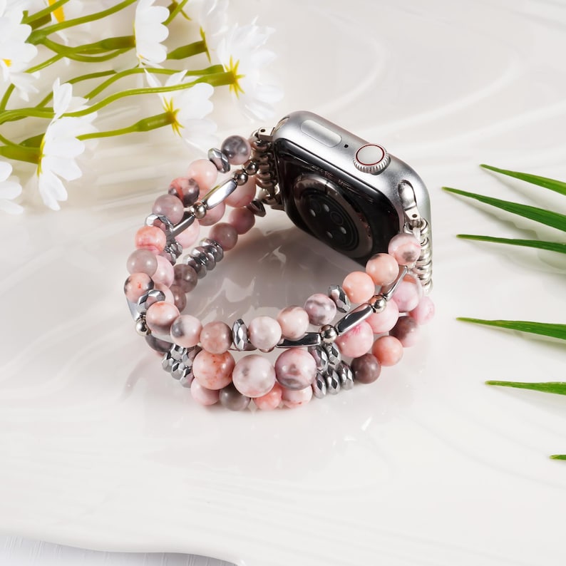 May include: A silver smartwatch with a pink and gray beaded band. The band is made of round beads in varying shades of pink and gray, connected by silver metal bars. The watch is resting on a white surface, with white flowers and green leaves in the background.