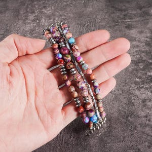 May include: A multi-strand bracelet featuring colorful, patterned beads in shades of purple, pink, blue, and brown. The beads are strung on a silver-toned metal wire. The bracelet is held in a hand.