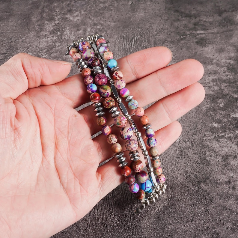 May include: A multi-strand bracelet featuring colorful, patterned beads in shades of purple, pink, blue, and brown. The beads are strung on a silver-toned metal wire. The bracelet is held in a hand.