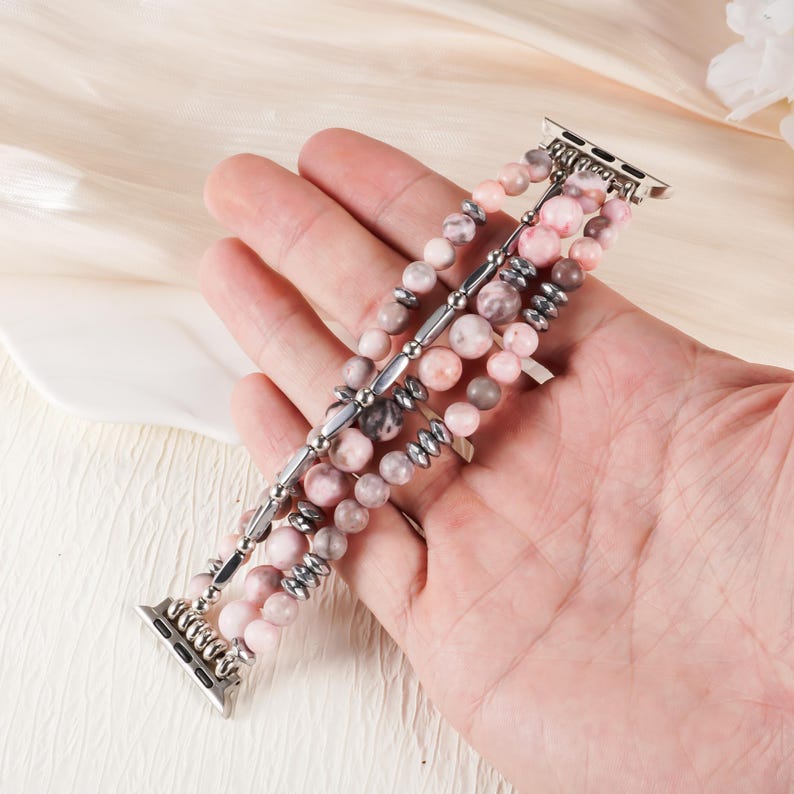 May include: A multi-strand Apple Watch band featuring pink and gray gemstone beads. The band has silver-colored metal connectors and accents. The beads vary in size and shade, creating a textured and visually appealing accessory.