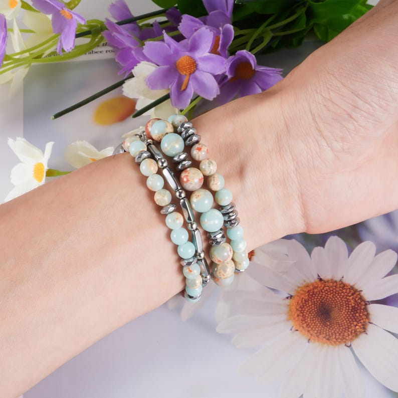 May include: A set of three beaded bracelets. The bracelets feature a mix of light blue, tan, and silver beads. The beads are arranged in a variety of sizes and shapes. The bracelets are worn on a wrist, with flowers in the background.