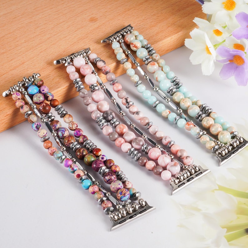 May include: Three beaded watch bands in shades of pink, brown, blue, and purple. Each band features multiple strands of round beads connected by silver-toned metal bars and clasps. The bands are arranged on a light wooden surface.