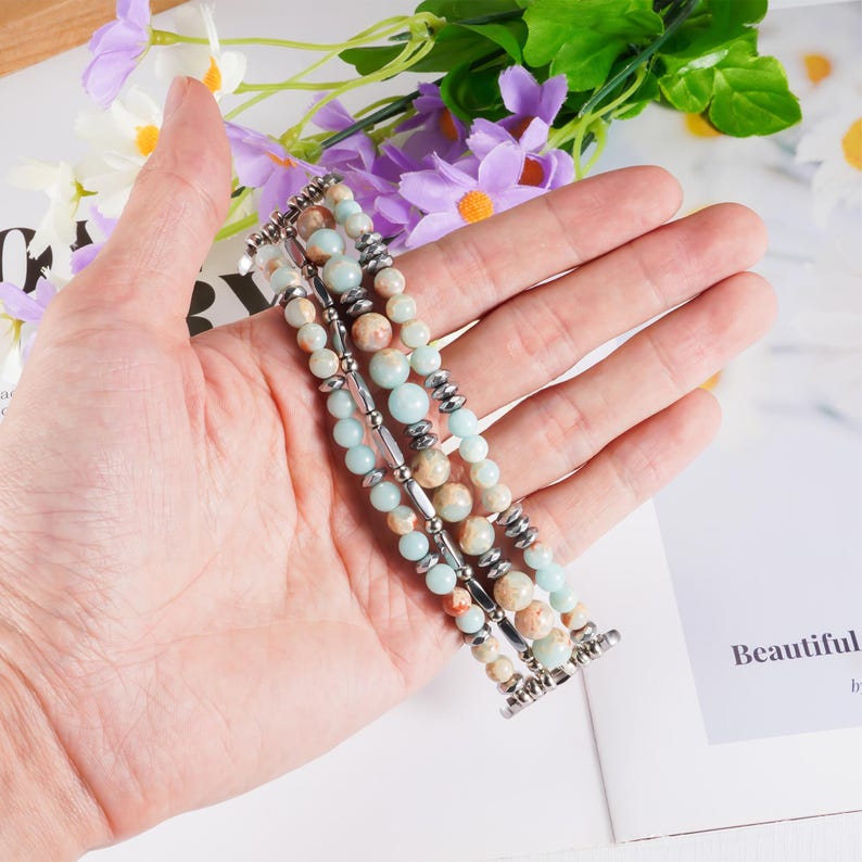 May include: A multi-strand bracelet featuring light blue, beige, and brown beads. The bracelet has silver-colored metal accents and clasps. The beads are of varying sizes and textures, creating a layered effect. The bracelet is held in a hand.