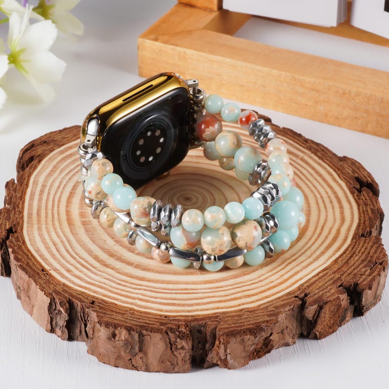 May include: A gold-colored smartwatch with a beaded band. The band features light blue, tan, and red-brown beads, interspersed with silver-colored metal accents. The watch and band are displayed on a wooden slice.