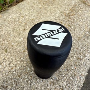 Può includere: Pomello del cambio nero con la scritta "Samurai" in bianco. Il pomello ha una parte superiore arrotondata e una forma cilindrica. Lo sfondo è una superficie strutturata di colore chiaro.