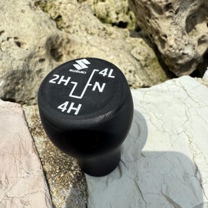 Può includere: Pomello del cambio Suzuki 4x4 nero con scritte bianche. Il pomello presenta il logo Suzuki e le marcature per 2H, 4H, N e 4L. Il pomello è realizzato in materiale nero opaco.