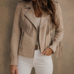 Puede incluir: Una chaqueta de textura de ante en color beige con largos flecos en el pecho y las mangas, decorada con tachuelas metálicas plateadas en las solapas, hombros y puños. La chaqueta incluye un cierre de cremallera frontal y dos bolsillos laterales.