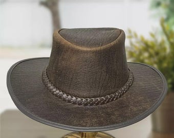 Può includere: Un cappello in pelle marrone scuro con una finitura testurizzata e vissuta. Il cappello presenta una corona pizzicata e una tesa larga leggermente curva. Una fascia in pelle intrecciata circonda la base della corona come dettaglio decorativo.