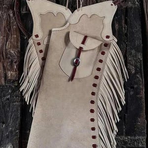 May include: Tan leather chaps with decorative fringe along the sides. The chaps have a pocket and are accented with dark red stitching and straps. The chaps are hanging against a dark wood background.